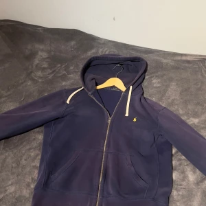 Marinblå huvtröja  Ralph Lauren - Säljer en snygg marinblå huvtröja från Ralph Lauren med dragkedja och broderad logga i gult. Storlek M men passar också S. Lite lila aktig i färgen då det är en äldre modell, men riktig skön tröja. 