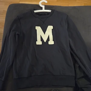 Mörkblå sweatshirt från Morris Stockholm - Säljer en mörkblå sweatshirt från Morris Stockholm med ett stort, vitt M på framsidan. Tröjan har långa ärmar och en rund halsringning. Perfekt för en avslappnad stil.
