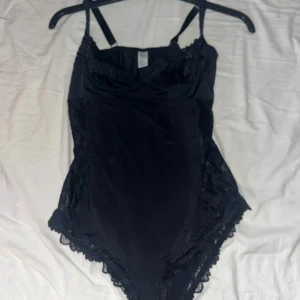 Svart spetsdetaljerad bodysuit - Elegant svart bodysuit med vackra spetsdetaljer. Den har justerbara axelband och en stilren design som passar perfekt. Oanvänd
