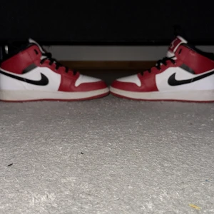Nike Air Jordans 1 mid chicago i rött och vitt - Säljer ett par klassiska Nike Air Jordans i rött, vitt och svart. Skorna har en ikonisk design med snörning och den välkända Nike-loggan på sidan. Perfekta för sneakerheads som vill ha en tidlös stil. De är helt äkta från restocks