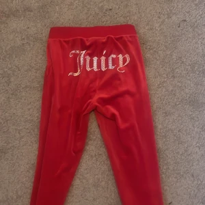 Röda mjukisbyxor från Juicy Couture - Snygga röda mjukisbyxor från Juicy Couture med glittrande text på baksidan. Byxorna har en bekväm passform med snörning i midjan och är perfekta för en avslappnad stil.