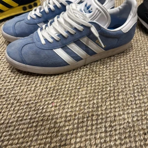 Blå Adidas Gazelle sneakers - Snygga blå Adidas Gazelle sneakers med vita ränder och snörning. Perfekta för en avslappnad stil. De har en klassisk design med en bekväm passform.
