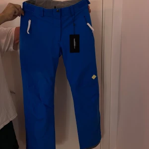 Blå skidbyxor från J.Lindeberg - Snygga blå skidbyxor från J.Lindeberg med dragkedja och knappar. De har en normal passform och är perfekta för en stilren look. Byxorna har även bälteshällor och en diskret logga baktill. Ny med prislapp 