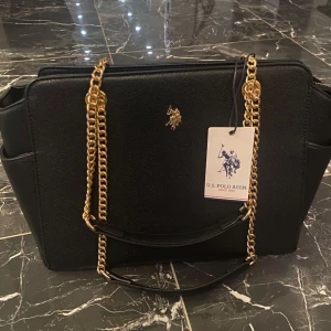 Svart axelväska från U.S. Polo Assn. - Snygg svart axelväska från U.S. Polo Assn. med guldfärgade kedjedetaljer och dragkedja. Väskan har ett rymligt innerfack med mönstrat foder och en praktisk innerficka med dragkedja och är helt ny.