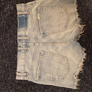Blå jeansshorts från Zara - Säljer ett par blå jeansshorts från Zara med slitna detaljer och fransiga kanter. Shortsen har en medelhög midja och klassisk femficksdesign. Perfekta för en avslappnad sommarlook! Köpta för 350kr och använda fåtal gånger pga för små!