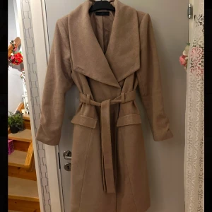 Beige kappa från Ellos - Elegant beige kappa från Ellos med knytskärp i midjan och bred krage. Kappan har långa ärmar och en stilren design som passar perfekt för kyligare dagar. Passar storlek M/L