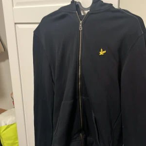 Svart huvtröja från Lyle & Scott - Säljer en svart huvtröja från Lyle & Scott med dragkedja och gul logotyp på bröstet. Tröjan har långa ärmar och en praktisk huva. Perfekt för en avslappnad stil.