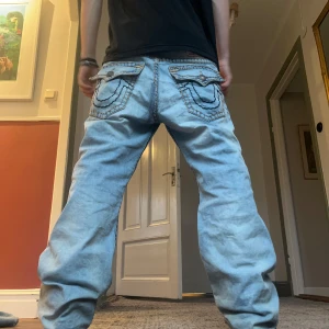 Ljusblå jeans från True Religion - Säljer ett par ljusblå jeans från True Religion i modellen Billy Relaxed Bootcut. De har snygga detaljer på bakfickorna och en avslappnad passform. Perfekta för en casual look!