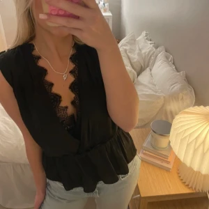 Svart ärmlös blus med spetsdetaljer - Snygg svart ärmlös blus från H&M med spetsdetaljer längs den djupa v-ringningen. Blusen har en omlott-design och en elastisk midja som ger en smickrande passform. Perfekt för en stilren look.