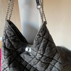 Grå quiltad axelväska med kedjedetaljer - Snygg grå axelväska med quiltat mönster och kedjedetaljer. Väskan har en stilren design med en praktisk axelrem och ett metallspänne framtill. Perfekt för att ge din outfit en trendig touch.
