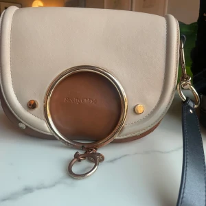 Beige och brun axelväska från SeeByChloé - Snygg axelväska i beige och brunt från SeeByChloé med en unik rund metallringdetalj på framsidan. Väskan har en justerbar axelrem och stängs med en magnetknapp. Perfekt för att ge din outfit en stilfull touch.