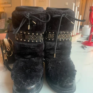 Svarta pälsboots från Love Moschino - Snygga svarta boots från Love Moschino med päls och dekorativa nitar. Skorna har snörning upptill och texten 'Love Moschino' runt skaftet. Perfekta för att hålla värmen med stil.