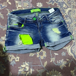 Blå jeansshorts med neon detaljer - Snygga blå jeansshorts från Miss Anna med coola slitningar och neon gröna detaljer. Den är lågmidjan och ganska fin att ha på sig på varma sommardagar. Den är XS men den passar ganska bra även på S