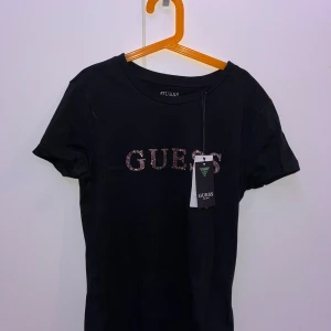 Svart t-shirt från Guess - Snygg svart t-shirt från Guess i ekologisk bomull. T-shirten har korta ärmar och ett glittrigt Guess-tryck på framsidan. 