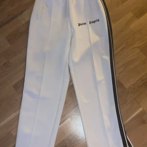 Vita byxor från Palm Angels - Snygga vita byxor från Palm Angels med svarta ränder längs sidorna. Perfekta för en stilren look. Storlek L men passar även M. Fickan är lite förstörd. Påse och lapp ingår vid köpet. 