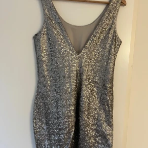 Silver paljett klänning  - Snygg glittrig silverklänning med djup v-ringning bak och ärmlös design. Perfekt för en festlig kväll med sitt glittrande material som fångar ljuset. Klänningen har en elegant passform som framhäver figuren.