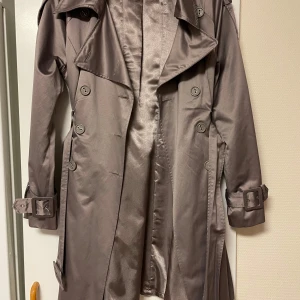 Grå trenchcoat från Derhy - Snygg grå glansig trenchcoat från Derhy i storlek M. Använd 2 gånger tidigare. Väldigt snygg och sitter fint. 