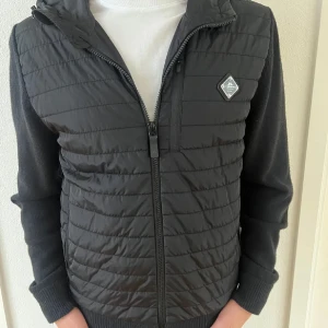 Svart quiltad jacka med dragkedja (J.Linderberg) - Snygg svart quiltad jacka med dragkedja och huva. Jackan har långa ärmar och en diskret logotyp på bröstet. Perfekt för kyligare dagar.