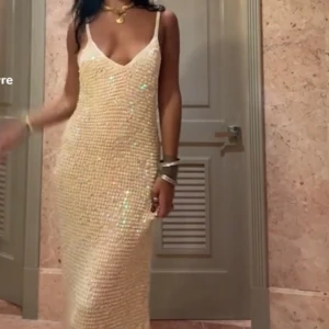 Glittrig vit långklänning - Snygg glittrig beige långklänning med smala axelband och djup v-ringning. Perfekt för en festlig kväll med sitt skimrande material som fångar ljuset vackert.