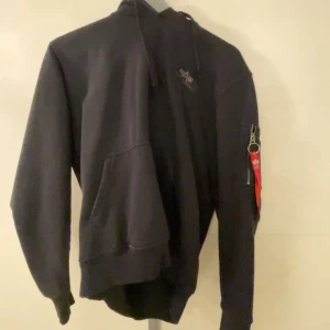 Svart hoodie från Alpha Industries - Snygg svart hoodie från Alpha Industries med stor logga på ryggen. Tröjan har en praktisk ficka framtill och en dragkedja på ärmen med en röd detalj. Perfekt för en casual look. ”””Dåliga bilder pga min kamera är sprucken””❤️