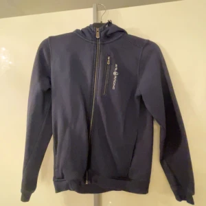 Mörkblå hoodie från sail racing - Snygg mörkblå hoodie från sail racing med dragkedja och huva. Perfekt för kyliga dagar. Den har en praktisk bröstficka med dragkedja och ribbade muddar vid ärmslut och nederkant. ”Dålig bild pga sprucken kamera”