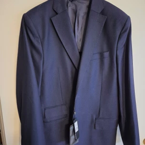 Mörkblå kavaj från J. Lindeberg - Snygg mörkblå kavaj från J. Lindeberg i slim fit. Tillverkad av exklusiv Super 120's ull från Loro Piana, vilket ger en lyxig känsla. Perfekt för eleganta tillfällen. Kavajen är ny och oanvänd med tag kvar.