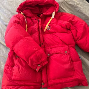 Röd dunjacka från Fjällräven - Säljer en röd dunjacka från Fjällräven med huva och dragkedja. Jackan har stora fickor med knappar och är perfekt för kalla dagar. Tillverkad i polyamid med fyllning av gåsdun. Säljer denna fjällräven jackan som är köpt på Vinted. Den har slitningar men ändå fin.