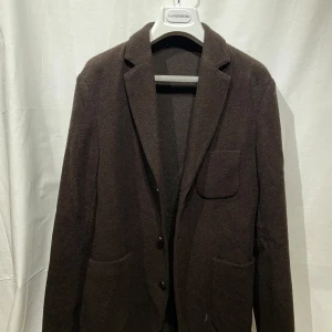 Kavaj från J.lindeberg - Snygg stickad brun kavaj från J.Lindeberg i ullmaterial. Kavajen har en klassisk design med knappar framtill och tre fickor. Perfekt för en stilren look. Knappt använd, modellen är 180cm