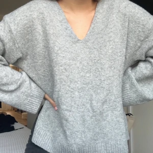 Grå stickad tröja med v-ringning - Mysig grå stickad tröja med v-ringad hals. Perfekt för kyligare dagar med sin avslappnade passform och ribbade kanter. En stilren och bekväm baströja för garderoben.