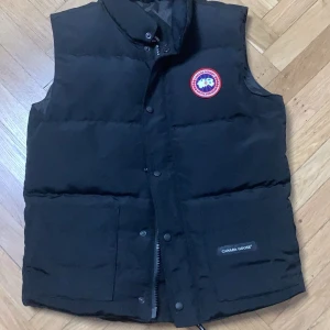 Svart dunväst från Canada Goose - Säljer en svart dunväst från Canada Goose med en broderad logotyp på bröstet. Västen har knappar och dragkedja framtill för extra värme och stil. Perfekt för kyliga dagar.