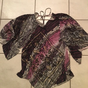 Mönstrad blus med knytband - Säljer en unik blus med ett färgglatt mönster i svart, rosa och beige. Blusen har korta ärmar och ett knytband vid halsen. Perfekt för att ge en färgklick till din garderob!