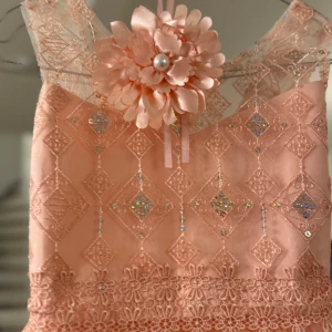 Persikofärgad klänning med spetsdetaljer - Charmig persikofärgad klänning med spetsdetaljer och en stor blomma framtill. Klänningen har ett glittrigt mönster och en luftig tyllkjol som ger en romantisk känsla.