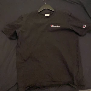 Svart t-shirt från Champion - Säljer en svart t-shirt från Champion med logotyp på bröstet och ärmen. Perfekt för en avslappnad stil. Tillverkad i mjukt material för extra komfort.