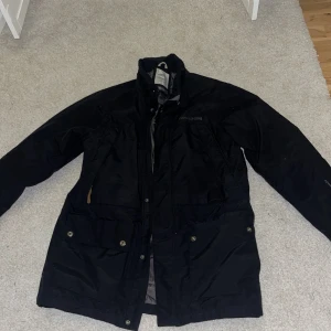 Svart jacka från Didriksons - Säljer en svart jacka från Didriksons med broderad logga på bröstet. Jackan har dragkedja och knappar framtill samt flera praktiska fickor. Perfekt för kyligare dagar.