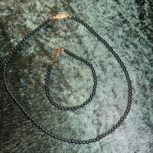 Svart pärlhalsband och armband - Elegant set med svart pärlhalsband och matchande armband. Båda smyckena har små, glänsande pärlor och dekorativa lås i guldton. Perfekt för att ge en stilfull touch till din outfit.