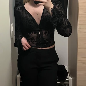 Svart blommig blus - Snygg svart blus med blommigt mönster och knappar framtill. Blusen har långa ärmar med volangdetaljer vid ärmslut. Oanvänd 