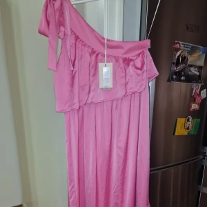 Rosa oneshoulder klänning - Snygg rosa oneshoulder klänning i satin med volangdetaljer. Perfekt för festliga tillfällen. Klänningen har en elegant och flytande design som ger en feminin look.