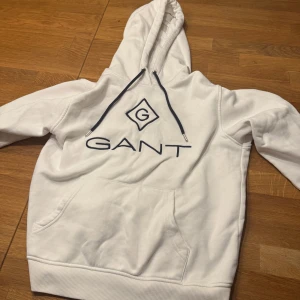 Vit hoodie från GANT - Säljer en stilren vit hoodie från GANT med stor logotyp på framsidan. Hoodien har en klassisk känguruficka och justerbar huva med snören. Perfekt för en avslappnad stil.