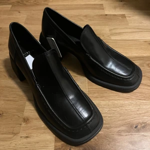 Svarta loafers från Vagabond 37 - Snygga svarta loafers från Vagabond i skinn med en stilren design. Skorna har en robust klack och en fyrkantig tå som ger en modern touch. Perfekta för en elegant look.
