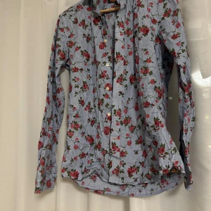 Blommig skjorta från Morris - Snygg skjorta från Morris med ett blommigt mönster i rött och grönt på en blå- och vitrandig bakgrund. Skjortan har långa ärmar och knappar framtill. 