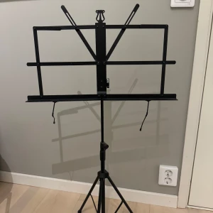 Svart notställ - Praktiskt och justerbart svart notställ perfekt för musiker. Stället har en stabil tripod-bas och justerbara armar för att hålla noterna på plats. Lätt att fälla ihop och ta med sig.
