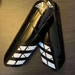Svarta benskydd från Adidas - Säljer ett par svarta benskydd från Adidas med vit logga och mönster. De kommer i en praktisk förvaringsväska och är perfekta för fotbollsträningen. Skydden är designade för att ge bra stöd och komfort.