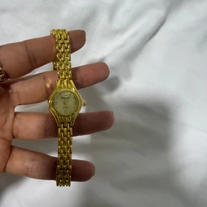 Guldfärgad klocka från Omega - Elegant guldfärgad klocka  med en klassisk design. Klockan har en rund urtavla och en länkarmband i guldton. Perfekt för att ge en lyxig touch till din stil.