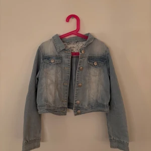 Blå jeansjacka från UNIQ - Säljer en snygg blå jeansjacka från UNIQ. Jackan har en klassisk design med knappar framtill och två bröstfickor. Perfekt för vår och sommar! Den har även spets längst ner på insidan av ärmarna så när man vecklar upp änden ser den extra snygg ut!  