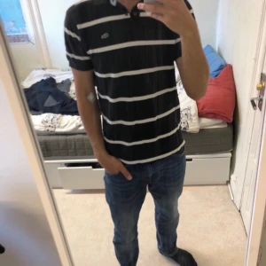 Svart och vit randig t-shirt från Lacoste - Snygg svart t-shirt med vita ränder från Lacoste. Den har en klassisk krage och korta ärmar, perfekt för en avslappnad stil. Den ikoniska krokodilloggan syns på bröstet.