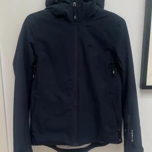 Mörkblå skidjacka från J.Lindeberg - Snygg mörkblå skidjacka från J.Lindeberg med dragkedja och huva. Jackan har långa ärmar och flera praktiska fickor med dragkedjor. Perfekt för utomhusaktiviteter och skyddar mot väder och vind. Endast använd 2 säsonger varav 1 vecka per säsong. Jättefint skick. Nypris 4000kr