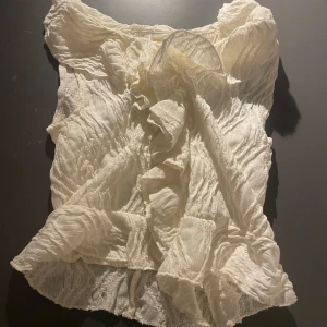 Vit blus med volangdetaljer - Elegant vit blus med volangdetaljer och en unik textur. Perfekt för att ge en romantisk touch till din outfit. Blusen är ärmlös och har en luftig passform.