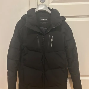 Svart stilren vinterjacka från Sail Racing - Snygg svart dunjacka från Sail Racing med huva och dragkedja. Perfekt för kyliga dagar. Jackan har en stilren design med logotyp på bröstet och ryggen samt vänstra armen. 