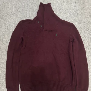 Vinröd stickad tröja från Polo Ralph Lauren - Snygg vinröd stickad tröja från Polo Ralph Lauren med hög krage och knappar. Perfekt för kyliga dagar med sin mysiga och stilrena design.