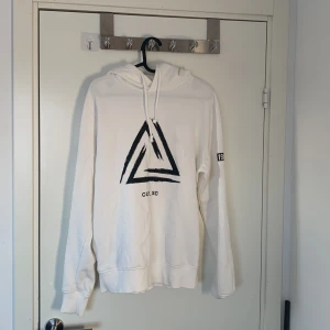 Vit hoodie från Jack & Jones - Säljer en vit hoodie från Jack & Jones med en stor svart triangel på framsidan och textdetaljer på ärmarna. Hoodien har en justerbar huva med snören och ribbade muddar vid ärmslut och nederkant. Perfekt för en avslappnad stil.